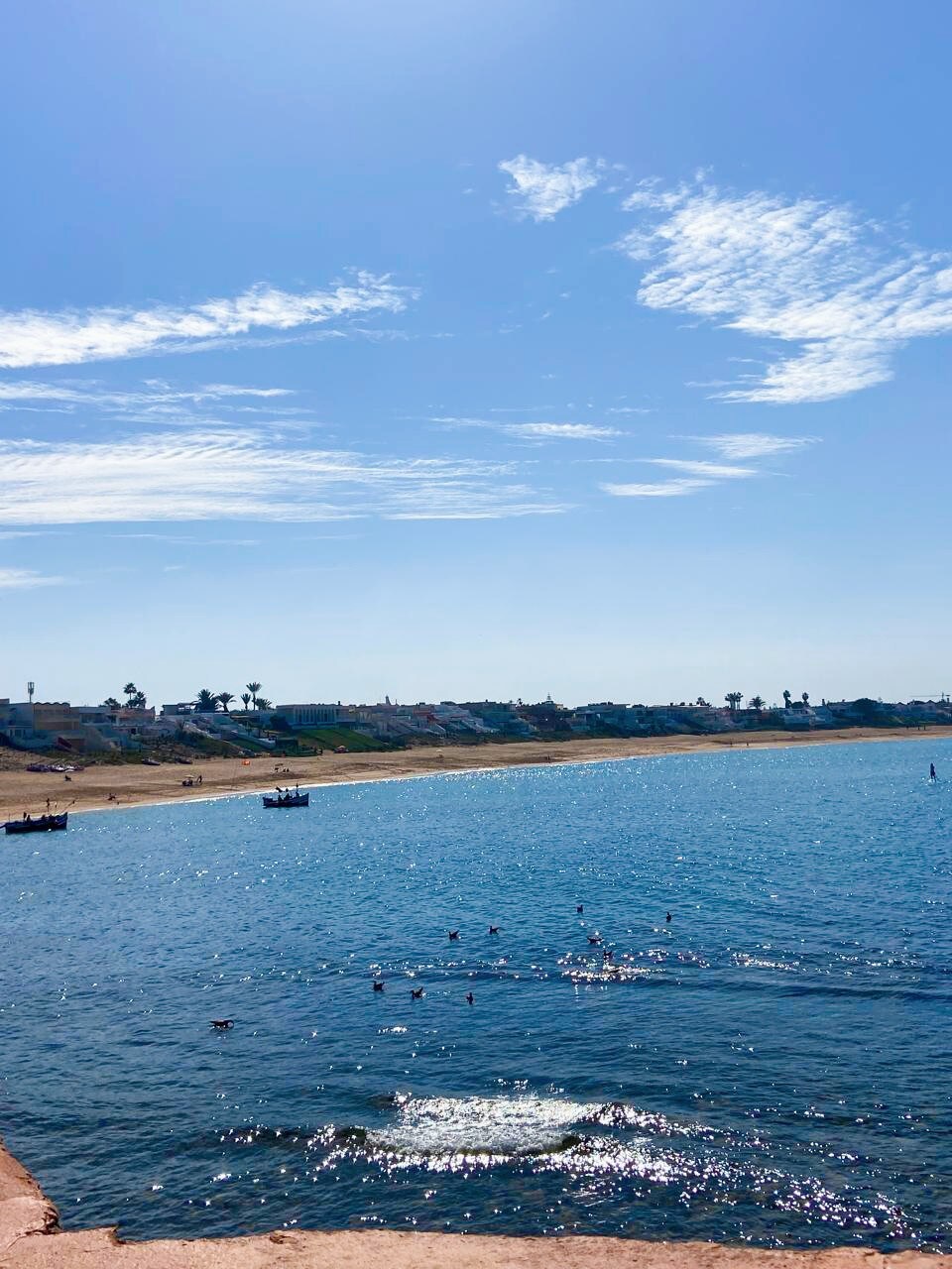 A serene beach scene is depicted, featuring clear blue waters gently lapping against the sandy shore. The sky is bright with a few scattered clouds, and several small boats can be seen in the distance, while people are swimming near the water's edge.