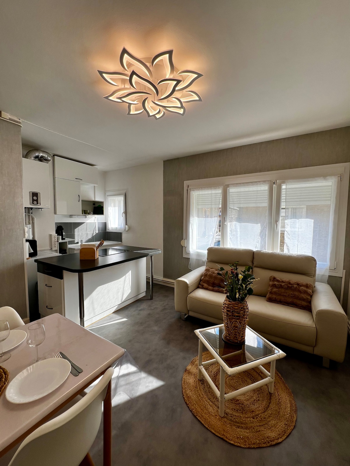 An open living area features a comfortable sofa facing a glass coffee table. Natural light floods through large windows, while a modern light fixture above adds an architectural element. The kitchenette is equipped with sleek cabinets and appliances, creating a cohesive and efficient space.