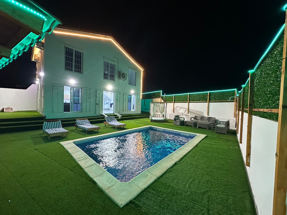 The outdoor area features a private pool surrounded by plush green grass. Several lounge chairs are arranged near the pool, alongside a seating area with comfortable sofas. Soft lighting outlines the house, creating a serene nighttime ambiance.