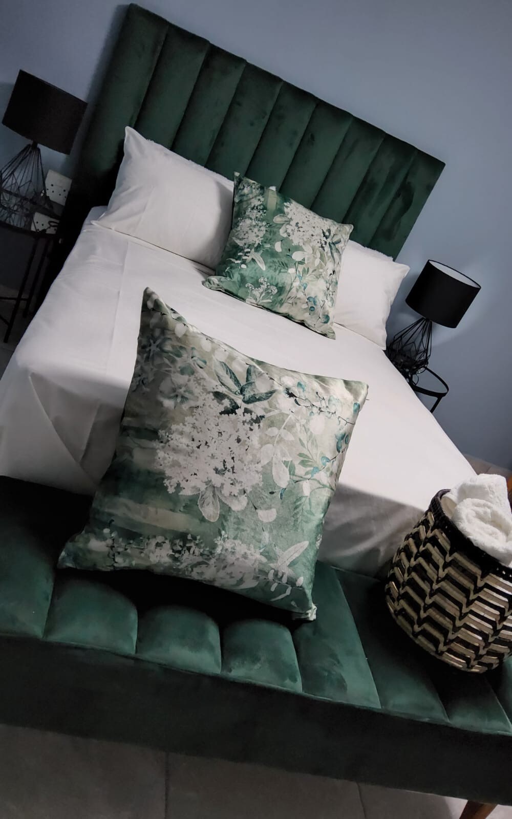 A stylish sleeping arrangement features a spacious bed with white linens and two decorative pillows in lush green floral patterns. On either side, modern bedside lamps add a touch of elegance. A patterned basket sits at the foot of the bed, enhancing the room's unique design.