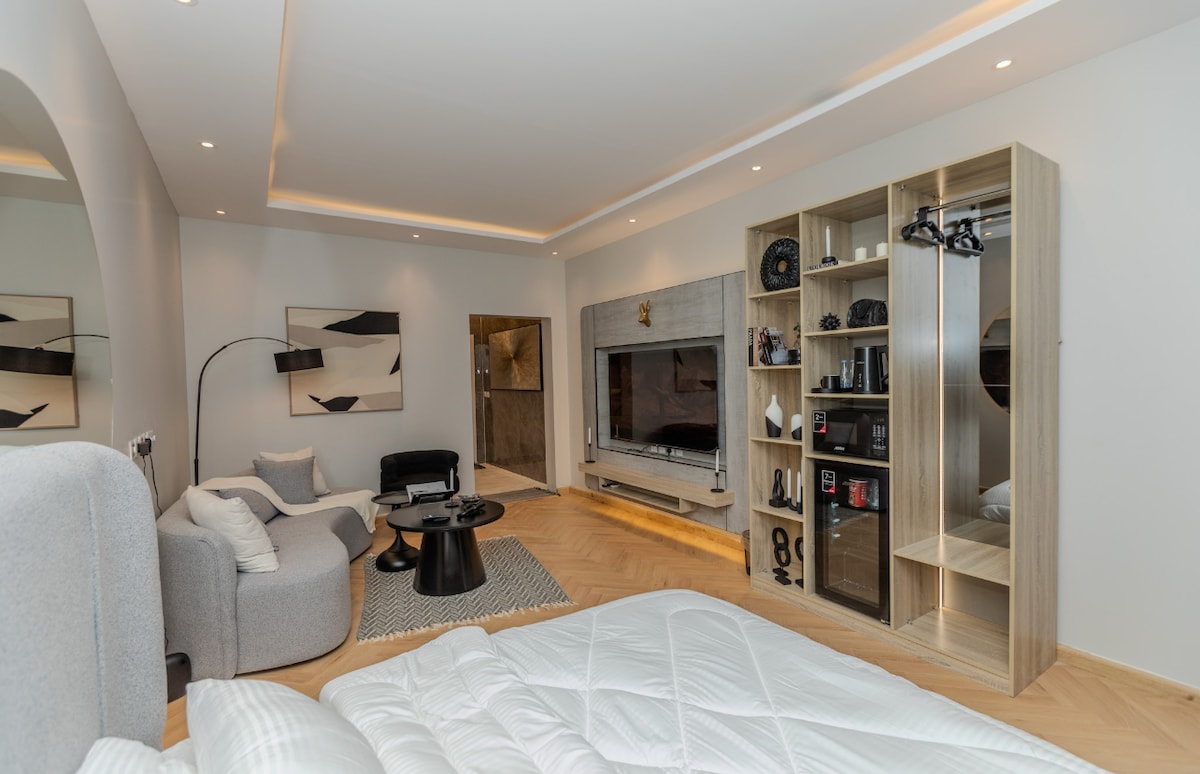 A modern living area is featured, showcasing a flat-screen television mounted on a light-colored wall. A comfortable sectional sofa and a round coffee table are arranged on a textured rug. A wooden entertainment unit displays decorative items and books, enhancing the space.
