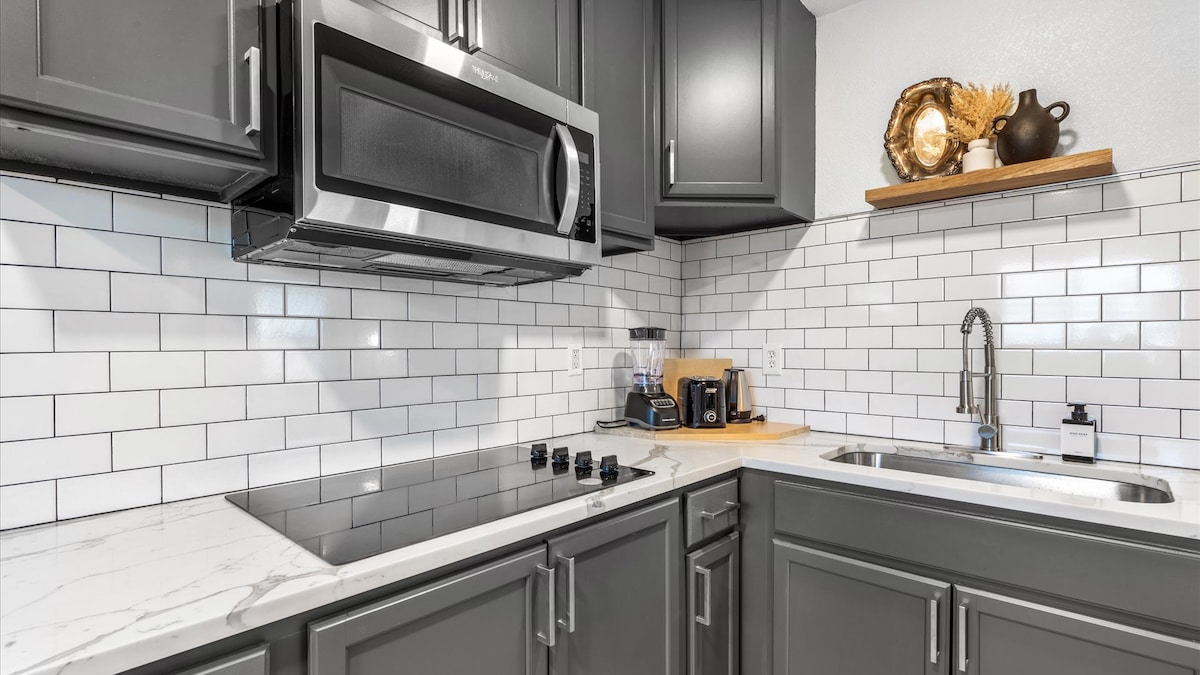 A modern kitchen features sleek dark cabinetry and a polished marble countertop. The arrangement includes a stainless steel microwave, a five-burner cooktop, and a contemporary faucet above a deep basin sink. A wooden shelf displays decorative items, enhancing the kitchen's functional design.