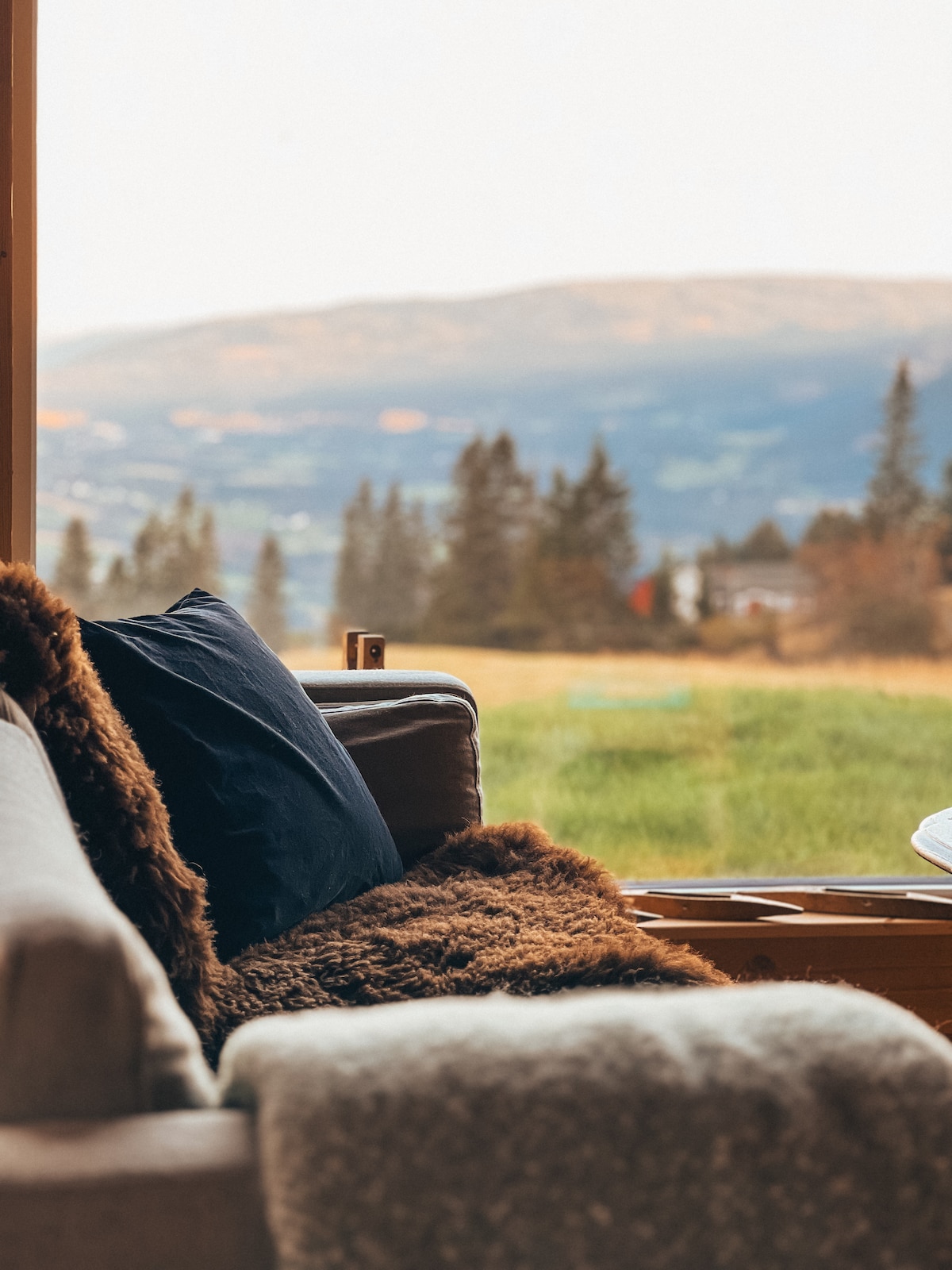 A cozy seating area features a soft, fur throw and cushions on a comfortable couch. Large windows provide a scenic view of the surrounding hills and fields, inviting natural light into the space.
