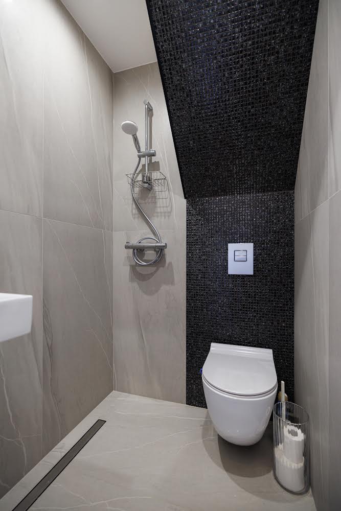 A modern bathroom features sleek, gray tiles with a glossy accent wall in black. The space includes a hanging shower head, a wall-mounted toilet, and a minimalist sink. A small waste basket is positioned beside the toilet for convenience.
