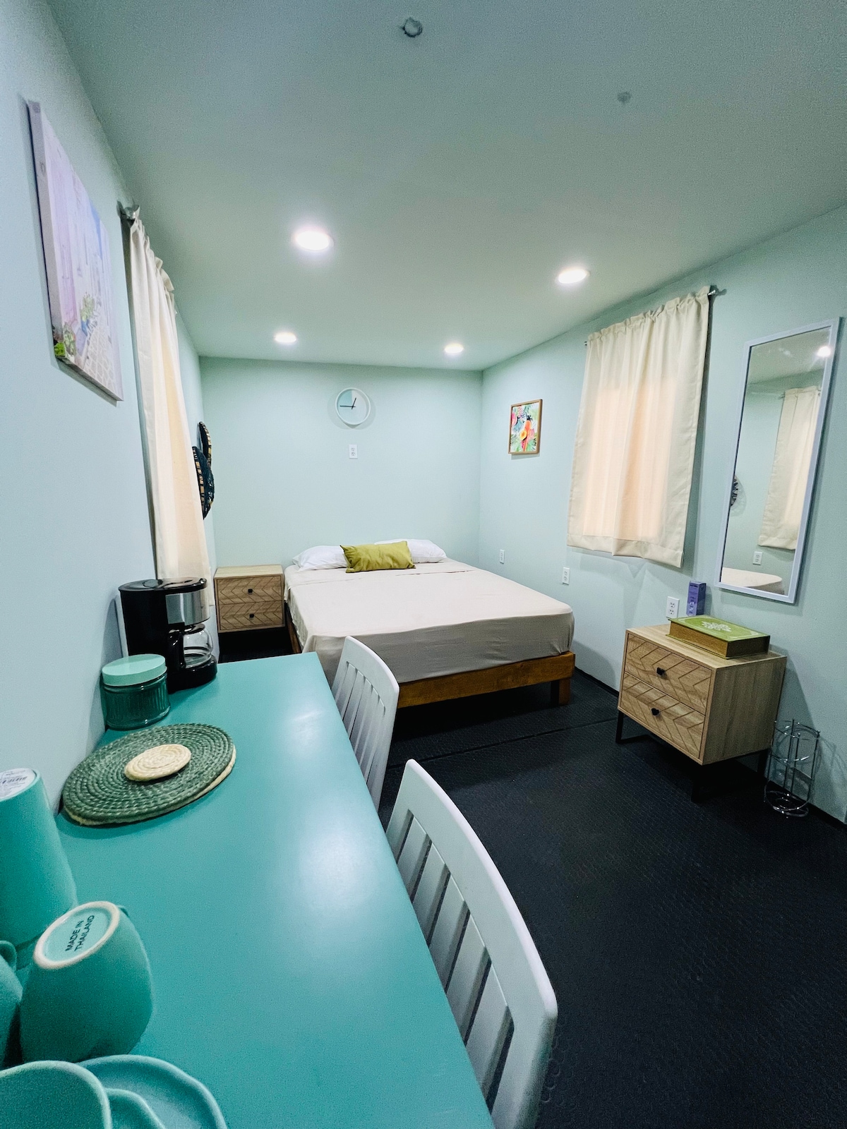 A serene bedroom features soft green walls and warm overhead lighting. A spacious bed is positioned against one wall, flanked by two nightstands. A small desk and chair occupy one corner, while a mirror reflects the light from the nearby window, framed with light curtains.