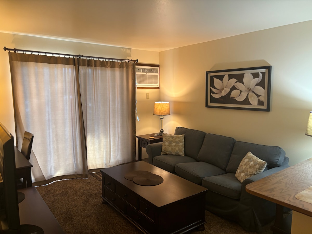 The living area features a cozy sofa positioned near a coffee table, complemented by two decorative pillows. A lamp illuminates the space next to a wall-mounted air conditioning unit. Soft curtains frame a window, and a piece of wall art adds a touch of elegance.