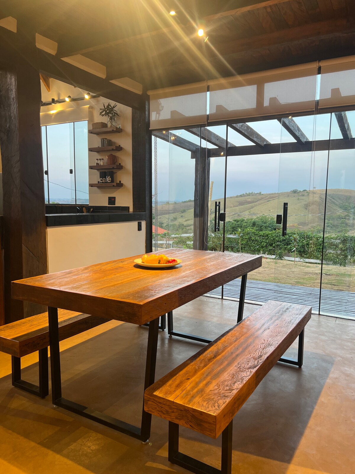 A wooden dining table with benches is positioned near glass doors that open to scenic views of the surrounding hills. Natural light floods the space, which features simple shelving on the wall for storage and additional decor.