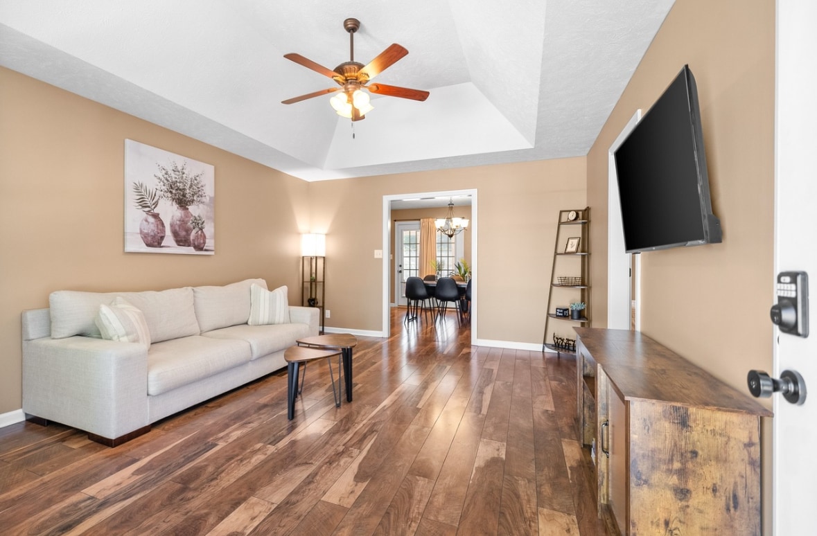A spacious living area features a light-colored sofa paired with a small, dark wood coffee table. A flat-screen television is mounted on the wall opposite the sofa. Natural light enters through a nearby doorway, highlighting the warm wood flooring.