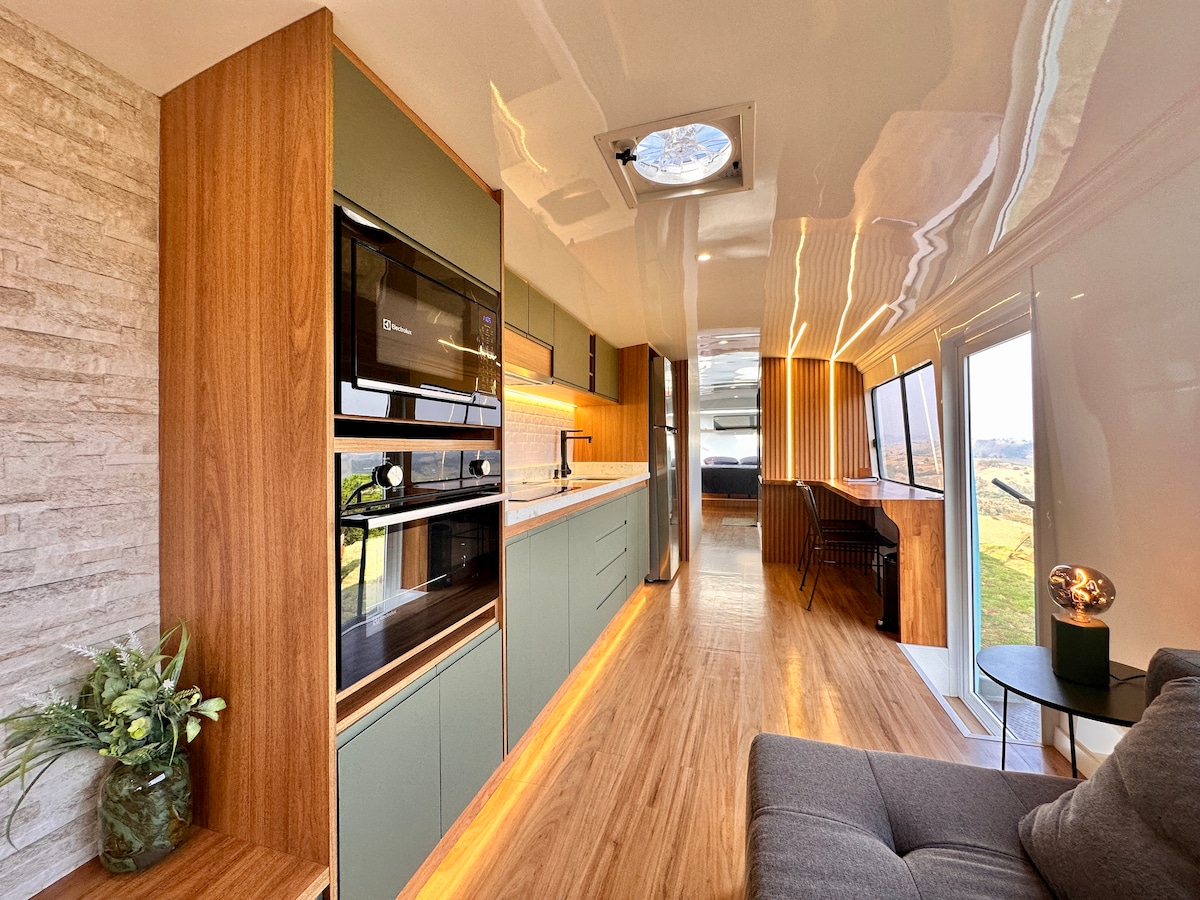 A modern interior space is presented, featuring sleek cabinetry and built-in appliances along a kitchen wall. Natural light floods the area through large windows, highlighting the wood paneling and smooth flooring. A cozy seating area is visible, enhancing the open, inviting layout.