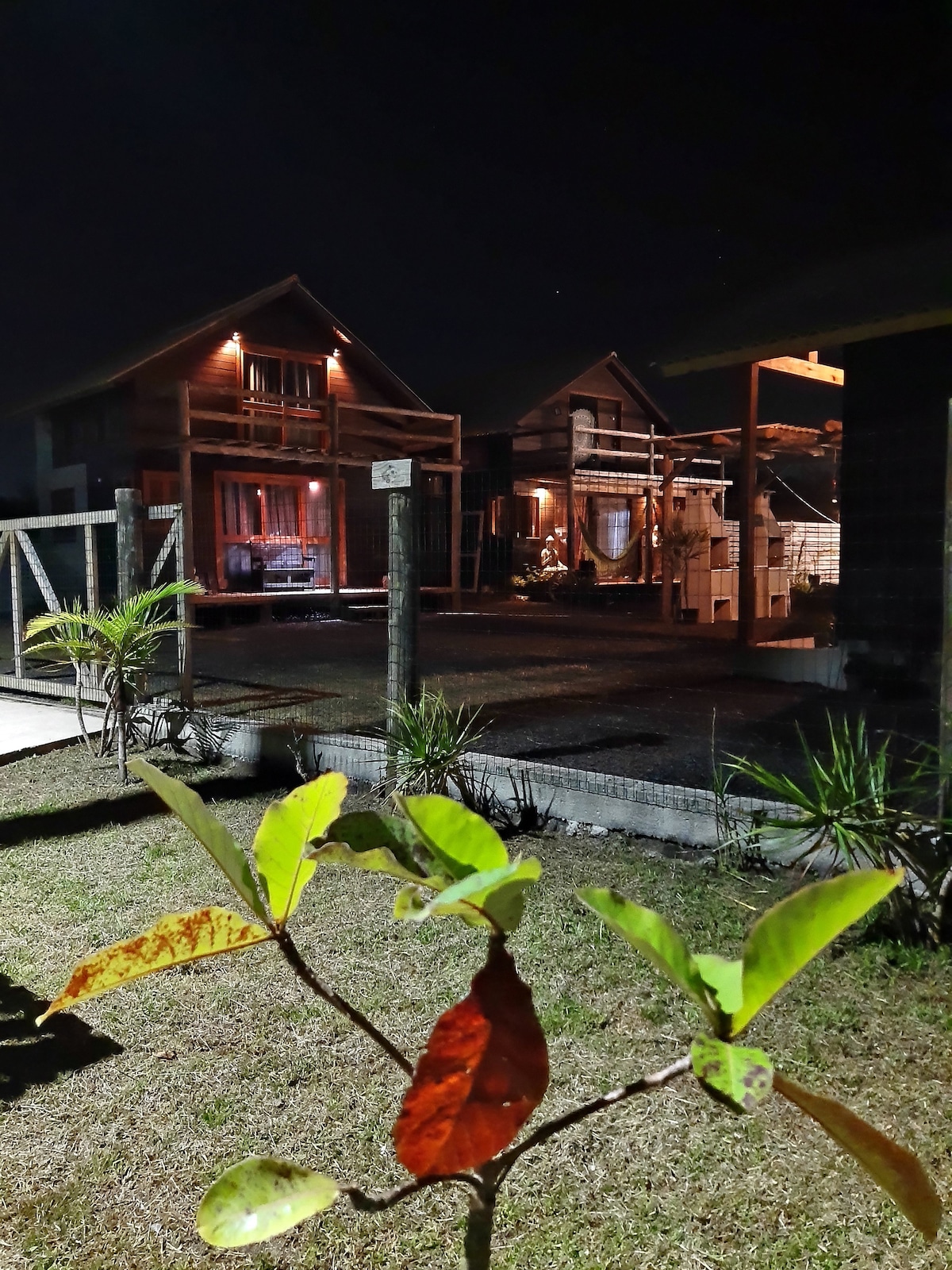 Two charming chalets are softly illuminated against the night sky, showcasing their inviting architecture. A well-maintained garden with plants is visible in the foreground, creating a peaceful ambiance. The layout suggests a cozy outdoor area for relaxation.