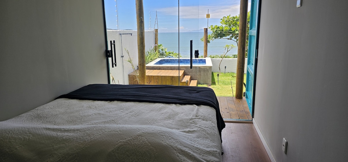 A spacious bedroom features a large bed dressed in light linens, with a wall of glass providing a view of the ocean and access to a private outdoor area. The tranquil scene includes a hot tub and greenery, enhancing the serene atmosphere.