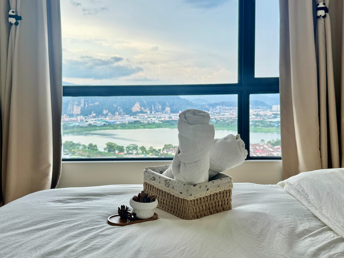 A cozy bedroom corner features a neatly made bed adorned with towels shaped like animals, presented in a woven basket. In the background, large windows offer a panoramic view of lush hills and scenic lakeside, inviting natural light into the space.