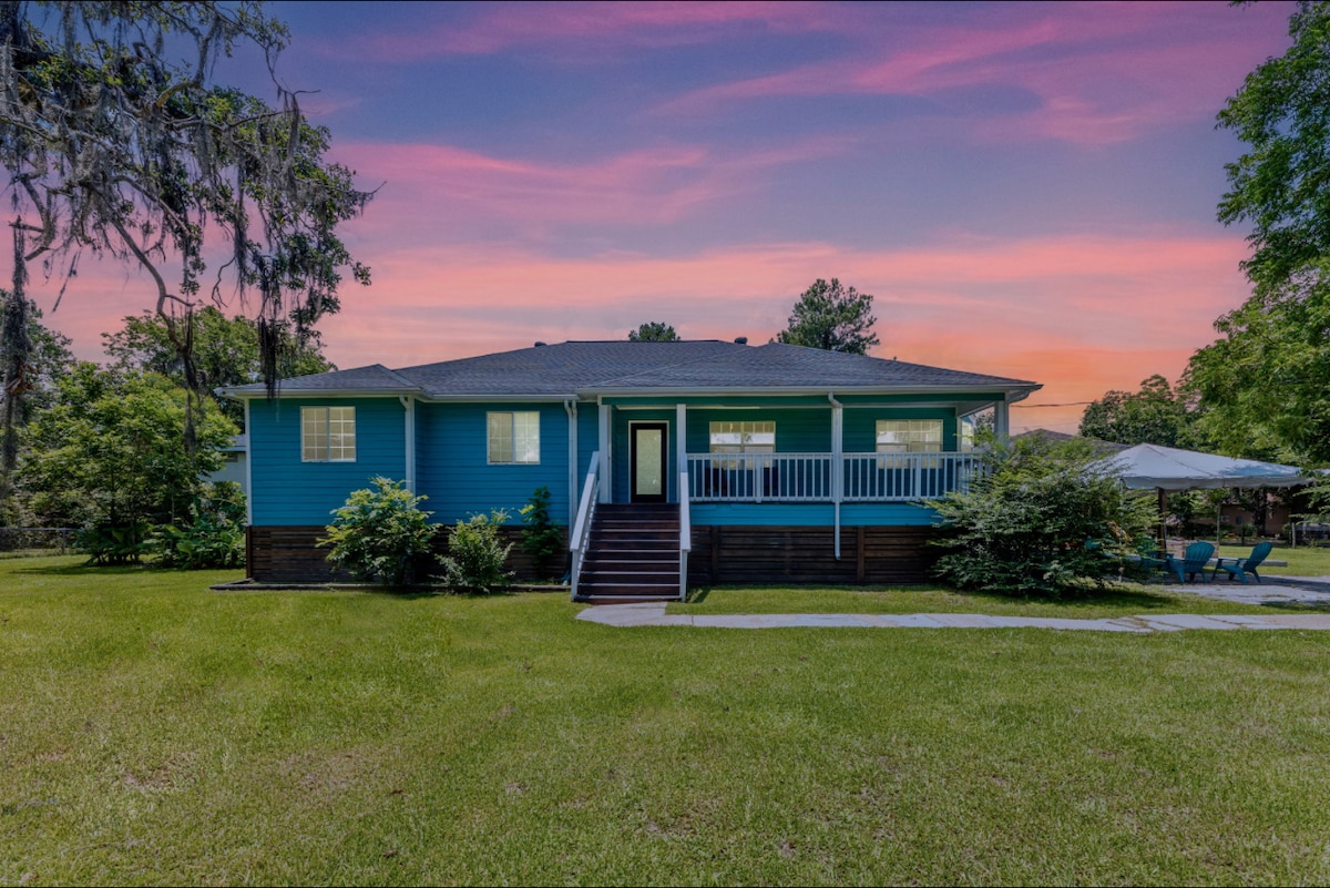 A single-story house with vibrant blue siding is set against a colorful sunset. The front porch features white railings and stairs leading to the entrance. Surrounding lush greenery and mature trees enhance the outdoor space, which includes a patio area with an umbrella.
