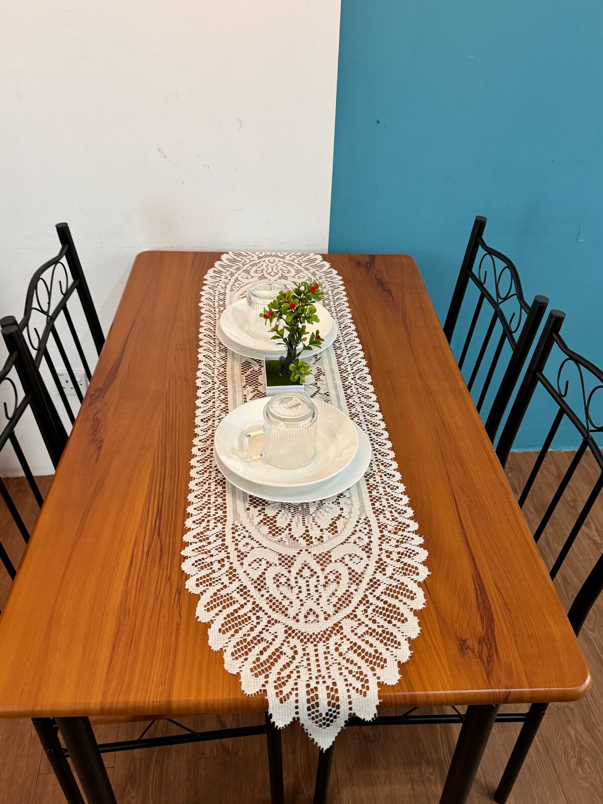 A wooden dining table is set for two, featuring a lace table runner that adds a touch of elegance. Each place setting includes a white plate and glass, accompanied by a small vase with fresh flowers, creating a welcoming dining atmosphere.
