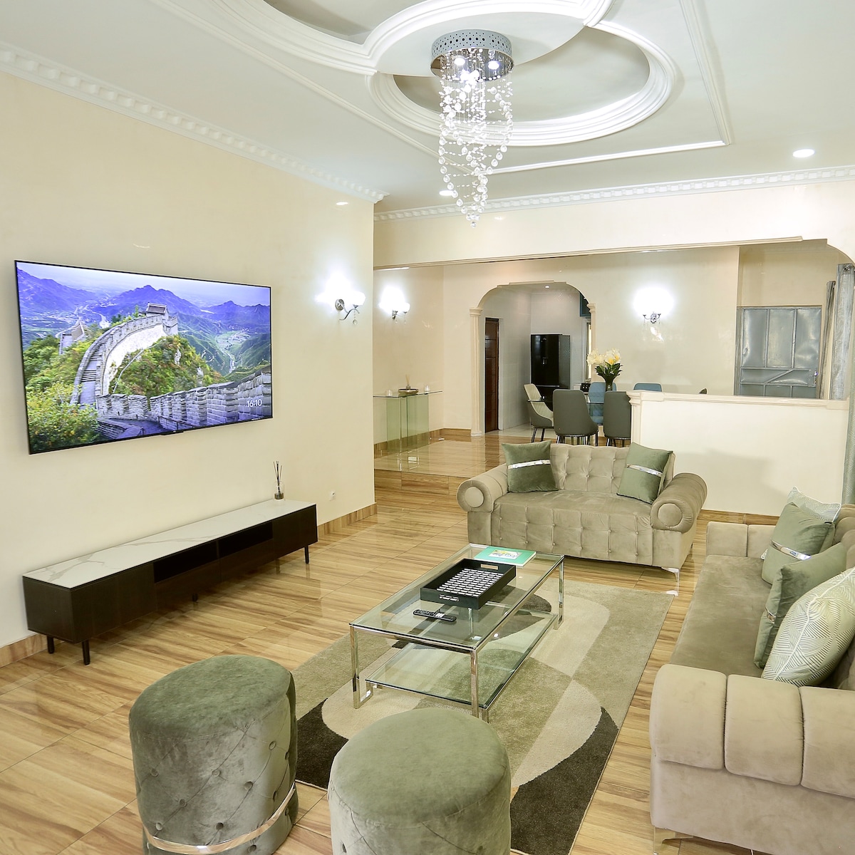 A modern living area is presented, featuring a large flat-screen TV mounted on a light wall. Elegant seating is arranged around a glass coffee table, complemented by two round ottomans. The room is illuminated by a stylish chandelier, enhancing the overall ambiance.