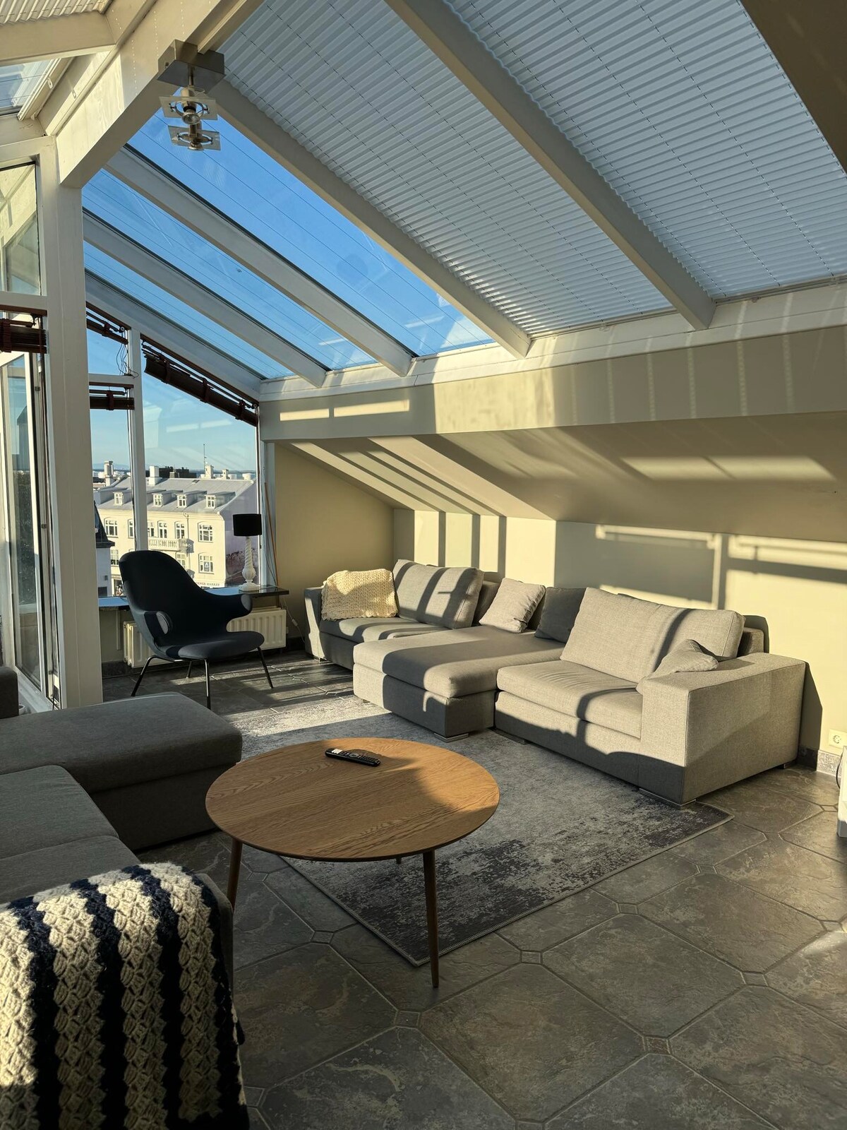 The living area features a large sectional sofa and an accent chair, with natural light streaming through the glass ceiling. A round coffee table sits in the center, surrounded by a textured rug, creating an inviting and bright space.