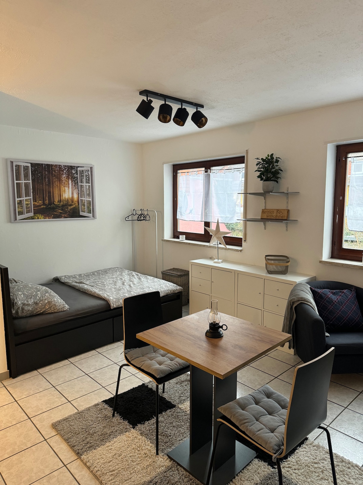 A well-lit studio apartment features a comfortable sleeping area with a spacious bed and a cozy seating arrangement around a wooden table. Two windows provide natural light, surrounded by light-colored walls and simple decor, including a wall-mounted artwork of a forest scene.