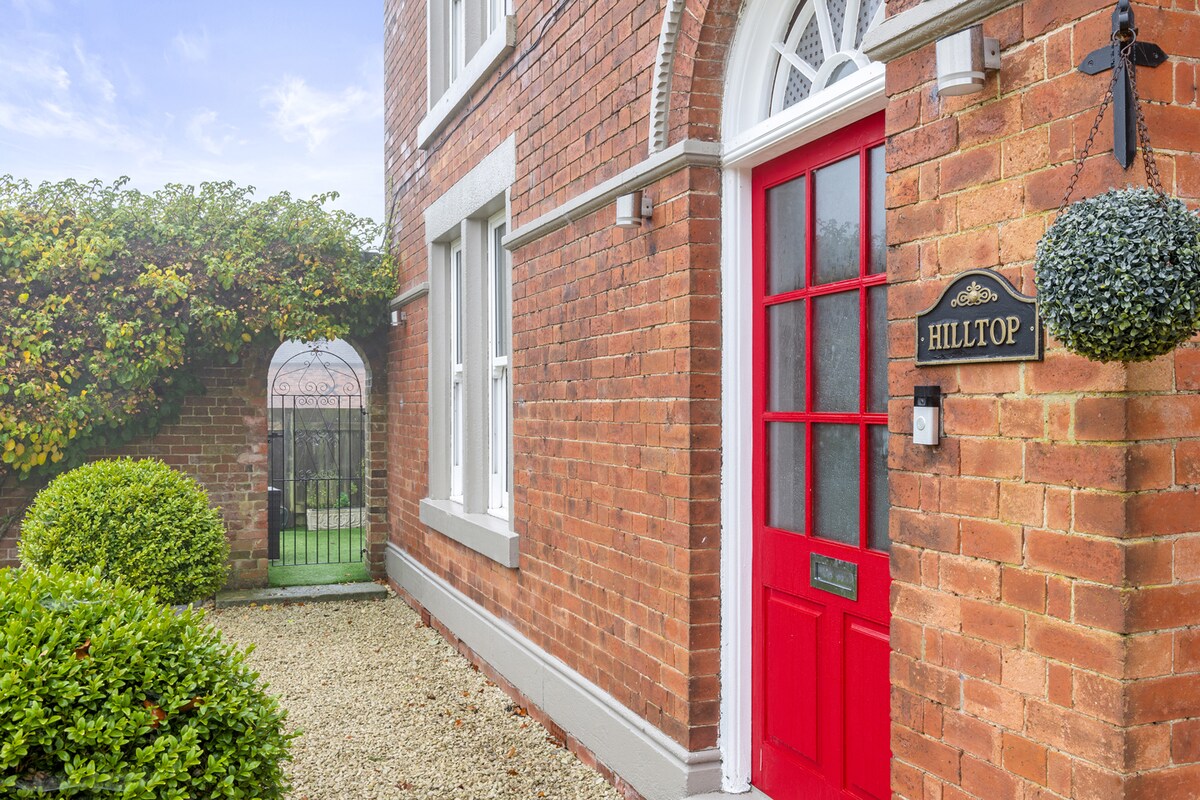A charming entrance is highlighted by a bright red door and a decorative nameplate. Lush greenery surrounds the entrance, with a well-maintained garden featuring neatly trimmed hedges and a gravel path. An elegant archway can be seen in the background, leading to a gated area.