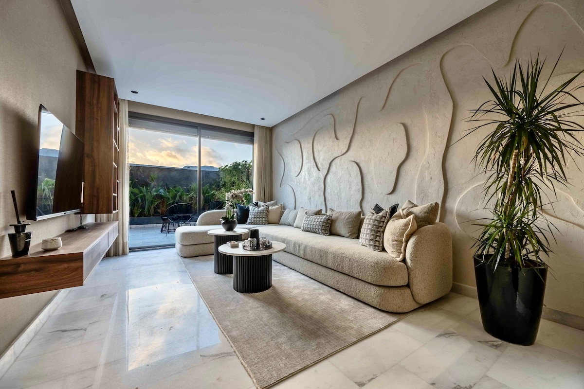 A spacious living area is revealed, featuring a comfortable sectional sofa adorned with neutral-toned cushions. A textured wall adds interest, while large glass doors offer a view of the outdoor space, allowing natural light to illuminate the marble flooring.