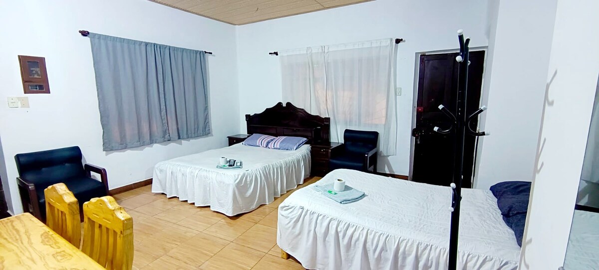 A spacious room features two beds covered with simple white linens. Light gray curtains frame the windows, while a wooden table and matching chairs are positioned nearby. Dark wood furniture adds warmth to the space, illuminated by soft indoor lighting.