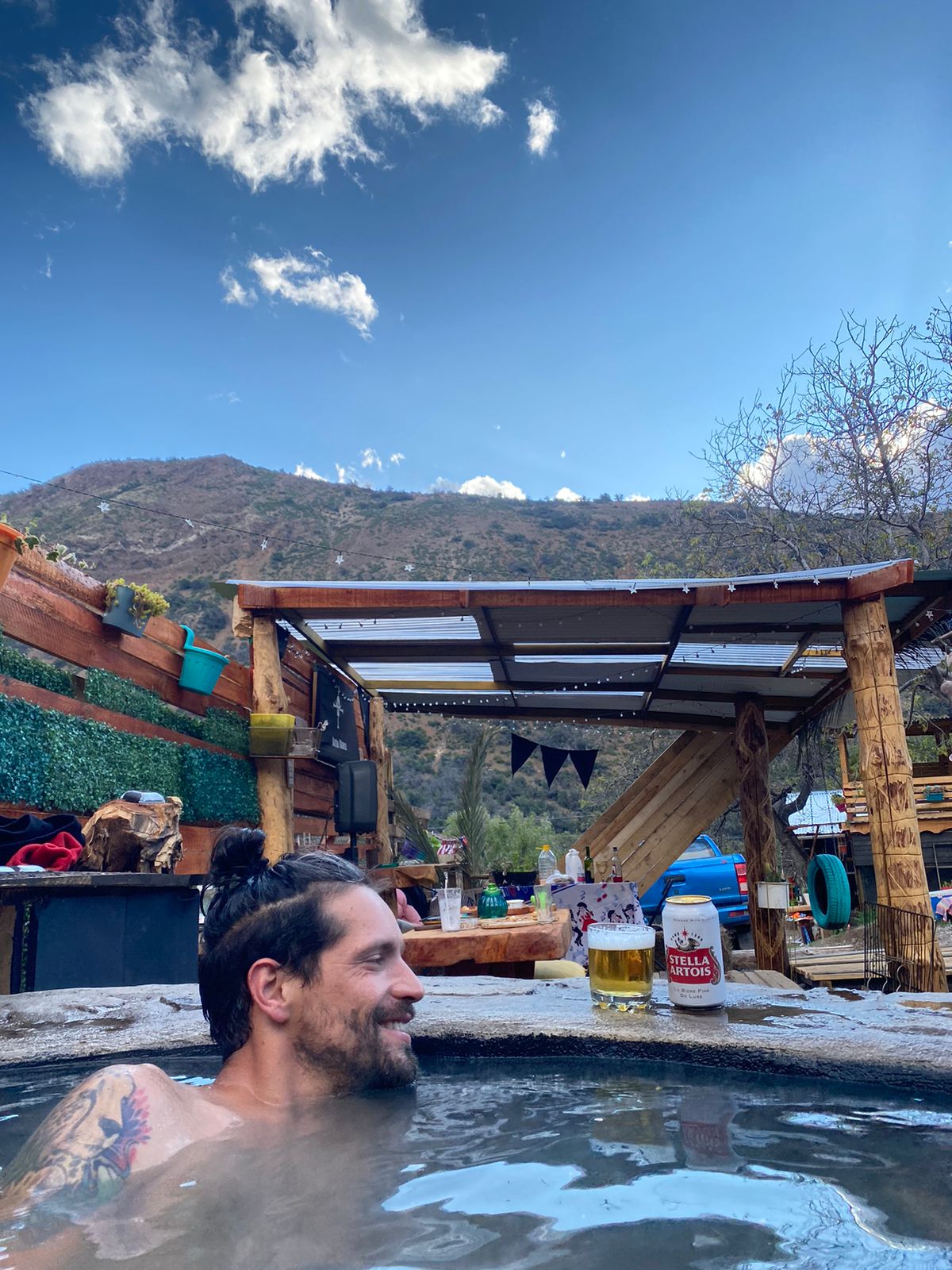 A hot tub is positioned outdoors, with steam rising softly against a backdrop of rolling mountains. Nearby, a rustic structure with a slanted roof and hanging decorations is visible, creating a relaxed atmosphere. Potted plants add a touch of greenery to the scene.