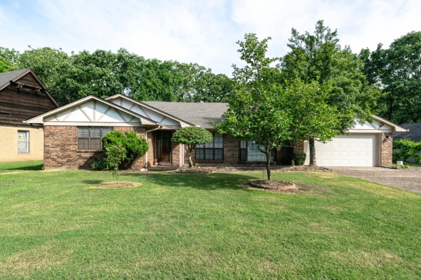 A single-story home is presented, featuring a combination of brick and wood siding. The property is set on a well-maintained lawn, accompanied by a circular driveway and a garage. Trees are positioned in the yard, offering a sense of greenery and tranquility.