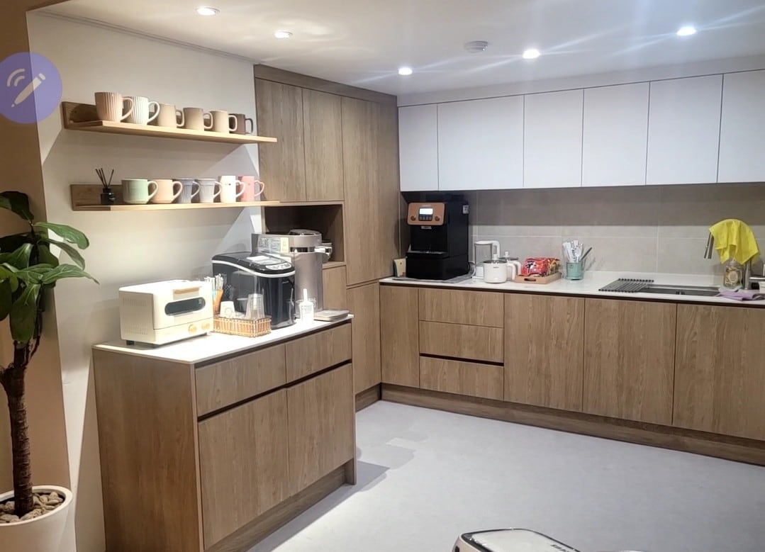 A modern kitchen area features wooden cabinetry and sleek countertops. A shelf displays various mugs, while a coffee maker and kitchen appliances are arranged for use. Soft overhead lighting creates an inviting atmosphere, complemented by a small potted plant in the corner.