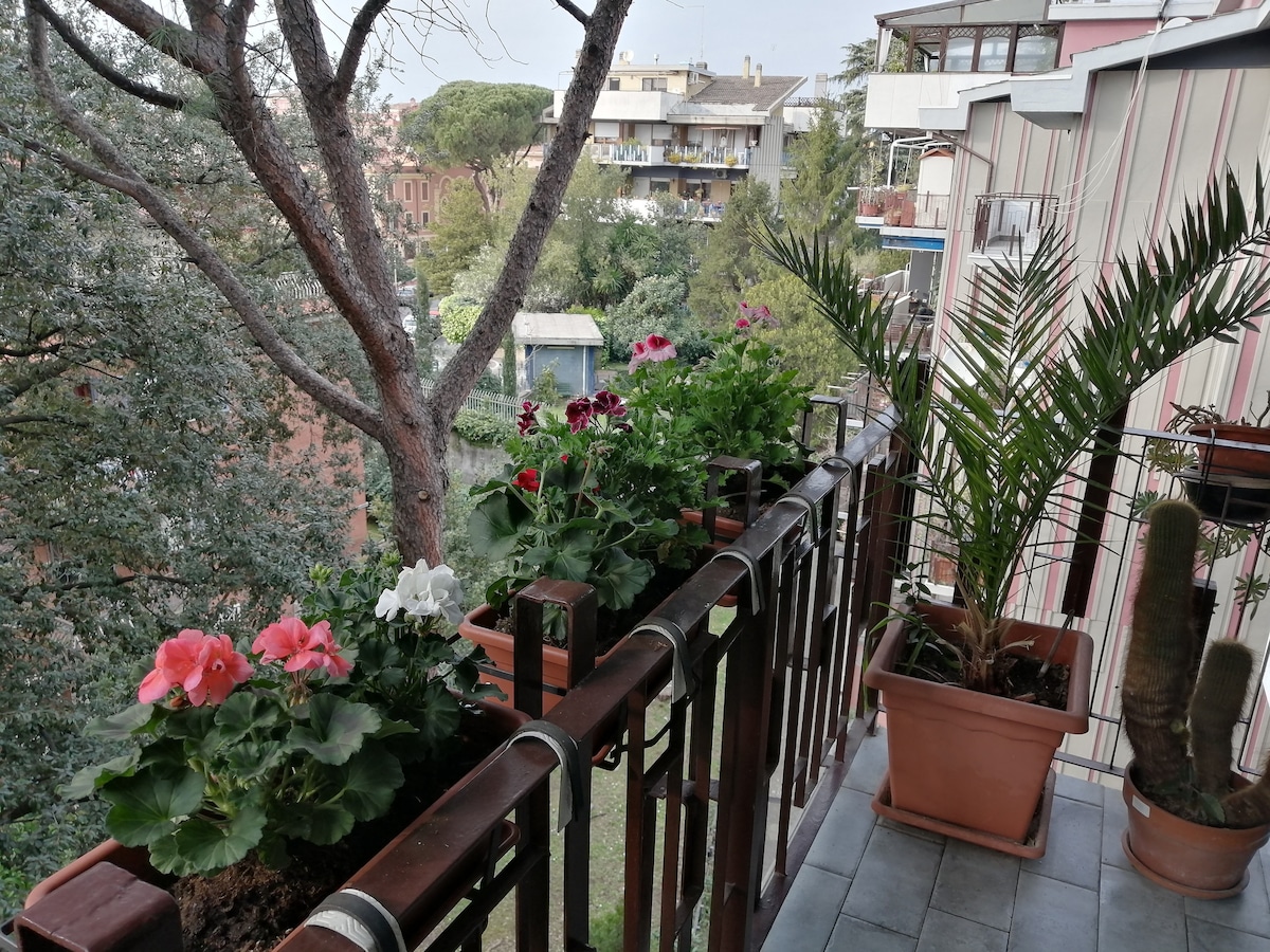 A balcony is adorned with several potted plants, including vibrant geraniums in various colors and a palm tree. The greenery creates a natural atmosphere, complemented by views of surrounding trees and buildings in the background.