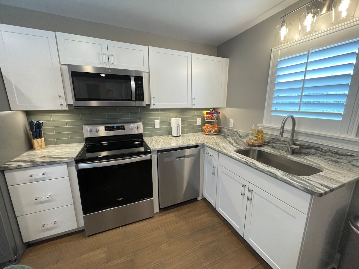 A modern kitchen features white cabinetry and a granite countertop. Stainless steel appliances include a microwave, oven, and dishwasher. Natural light streams in through a window adorned with shutters, illuminating the well-organized space. Fresh fruit is displayed in a bowl on the counter.