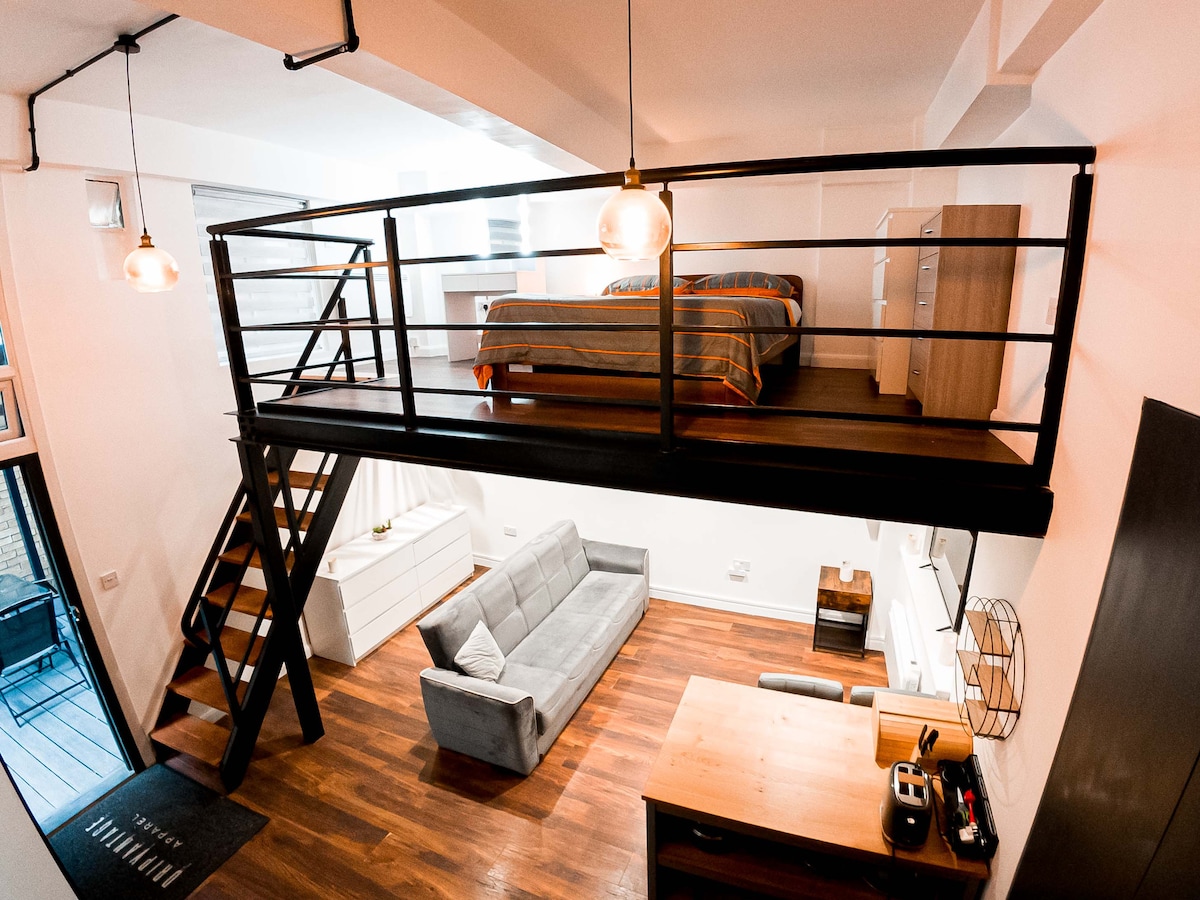 An aerial view of the studio apartment showcases a lofted sleeping area with a double-size bed, adorned with patterned bedding. A comfortable sofa is positioned below, alongside a wooden coffee table and a small desk, all set against warm wooden flooring.