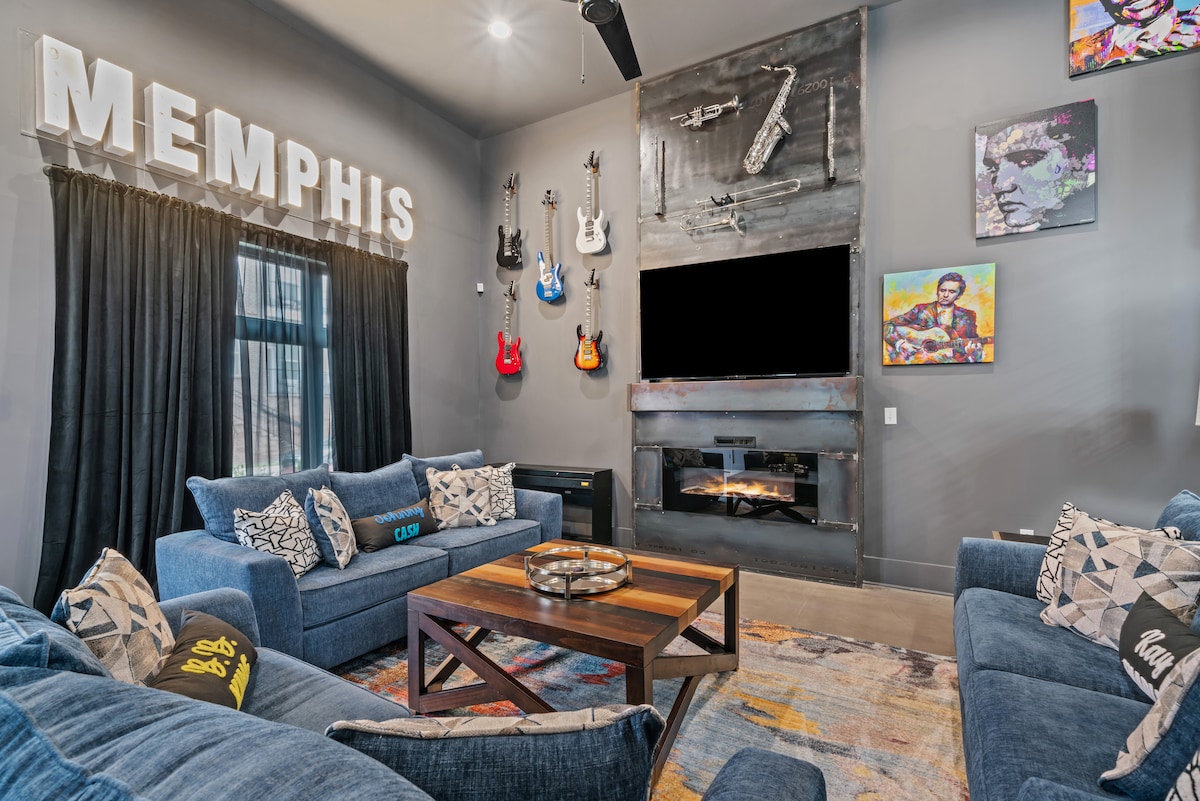 The living room features a modern design with dark gray walls and plush blue seating. The word 'MEMPHIS' is prominently displayed on the wall. Five guitars are arranged artistically, and a flat-screen TV is mounted above a stylish fireplace. A coffee table rests on a vibrant area rug.