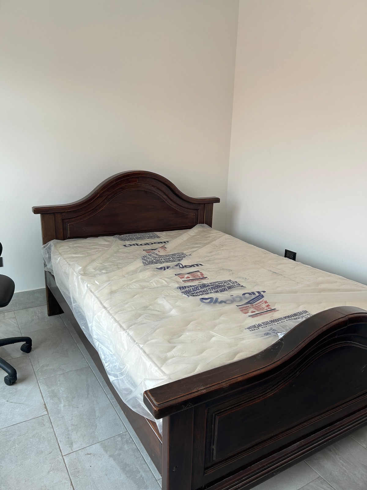 A spacious bed with a wooden frame is featured in the room, topped with a new mattress still in protective covering. A black office chair is positioned nearby on the tile flooring, contributing to the room's functionality.