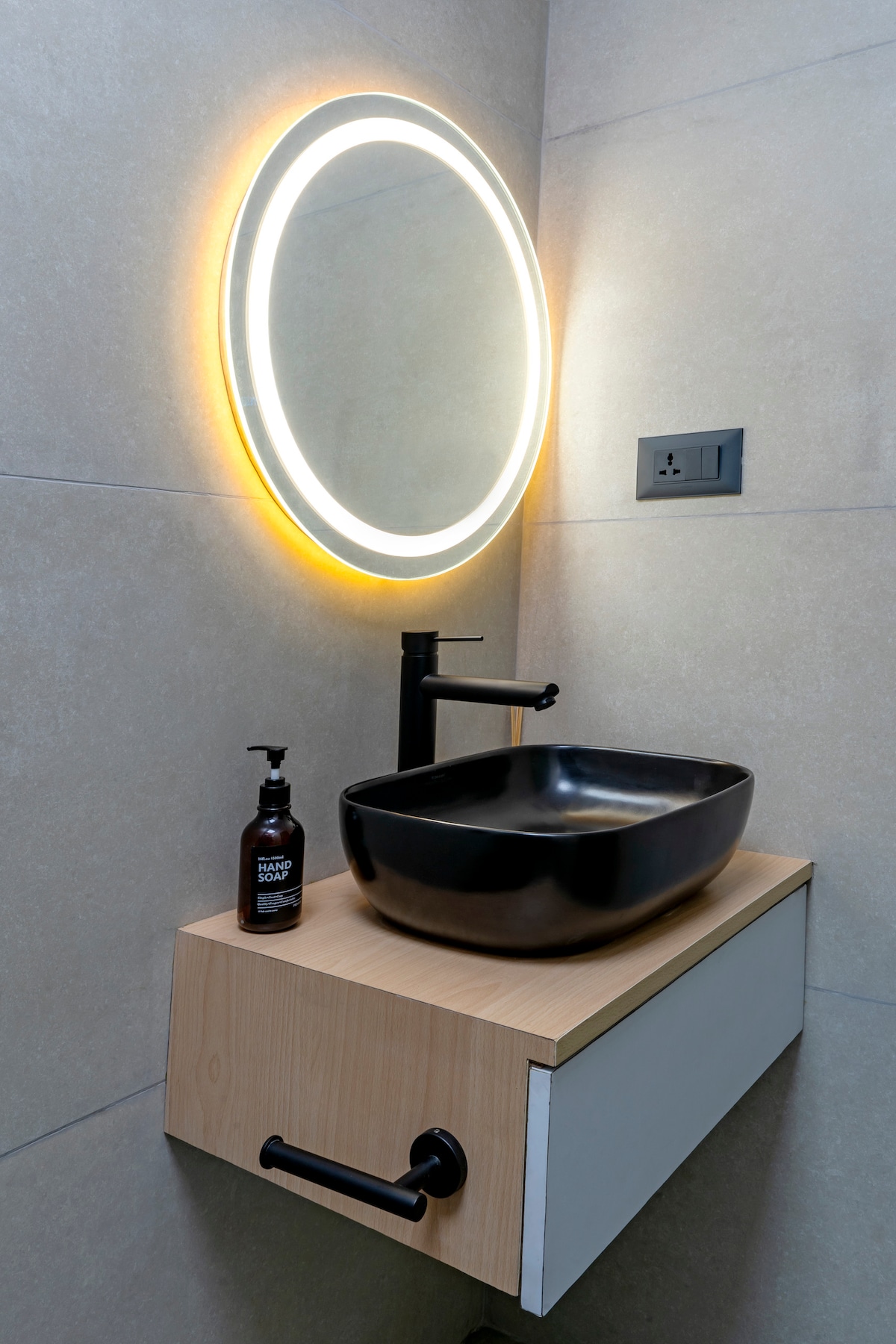 A modern bathroom sink area features a sleek black bowl sink integrated into a light wood countertop. A backlit circular mirror provides soft illumination, accentuating the clean lines of the space. A dark bottle of premium hand soap is positioned nearby for convenience.