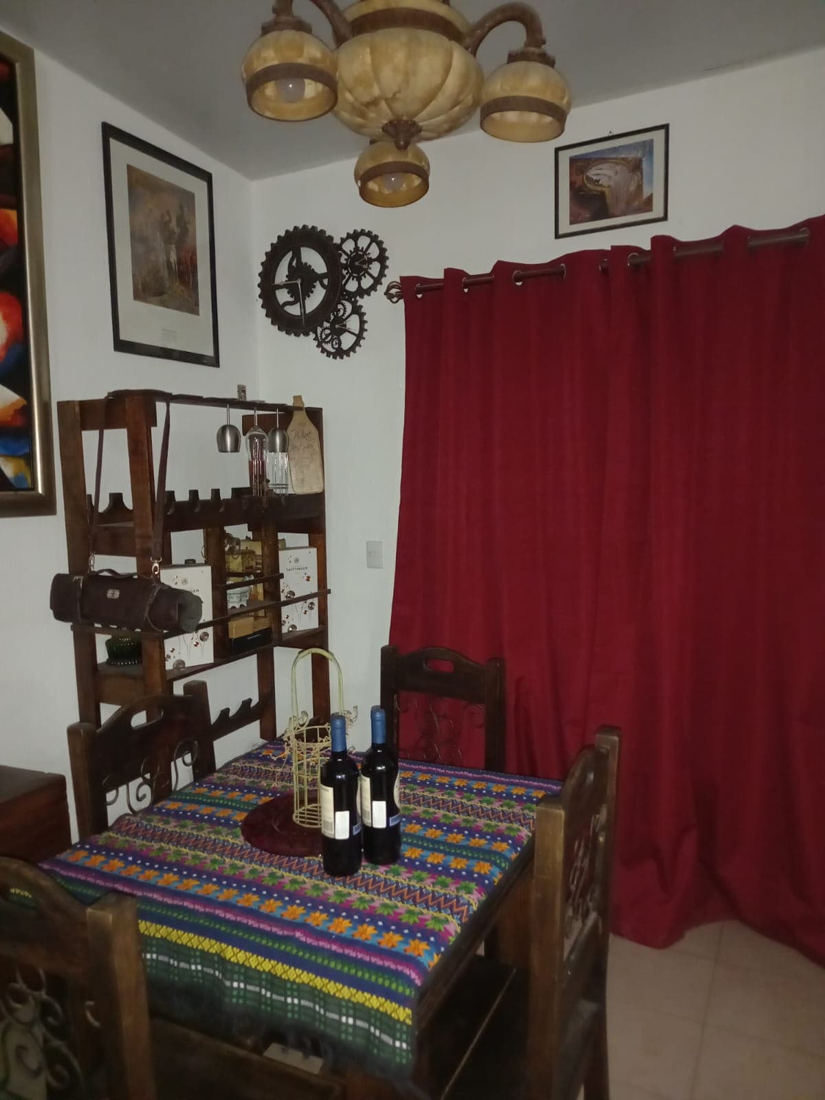 A dining area features a wooden table set with several wine bottles. Colorful tablecloths and decorative items are displayed alongside the wall, while a striking red curtain provides a bold accent. Unique art pieces are hung, adding character to the space.