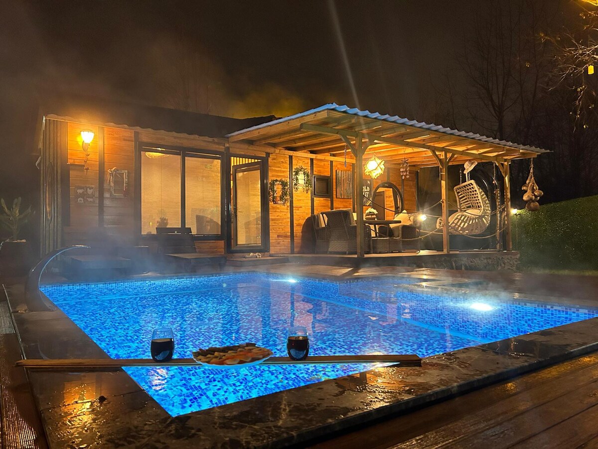 A heated pool glows with blue lighting under a wooden shelter. Steam rises gently into the night air, enhancing the cozy atmosphere. The wooden structure features large glass windows, providing views of a welcoming interior furnished with comfortable seating.
