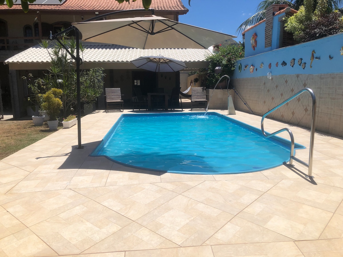 A clear blue pool is framed by a spacious tiled deck, featuring lounge chairs and large umbrellas for shade. Nearby, lush greenery is visible alongside a beautifully textured wall, contributing to a serene outdoor setting that invites relaxation.