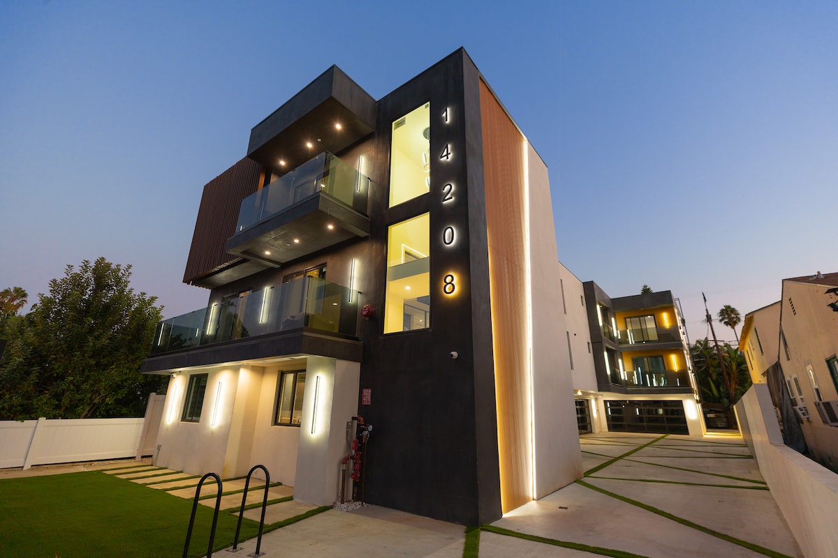A modern three-story building is showcased under a twilight sky, featuring a combination of sleek materials and glass balconies. The structure's illuminated numbers highlight its address, while a manicured lawn and pathways contribute to the inviting exterior.