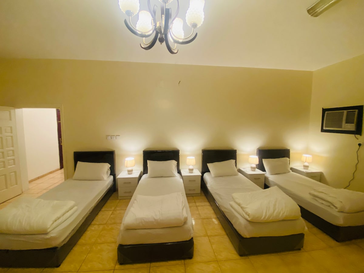 A spacious room is arranged with five single beds, each paired with a bedside table and lamp. Soft white linens are neatly laid on each bed, and a ceiling light fixture adds illumination to the area. The light-colored walls enhance the overall brightness.