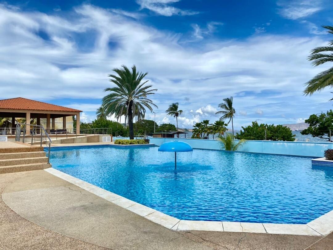 A large outdoor swimming pool is framed by a sunlit landscape, featuring palm trees swaying gently in the breeze. A shaded area with a roof is visible nearby. A bright blue umbrella stands in the center of the pool, complementing the clear sky above.