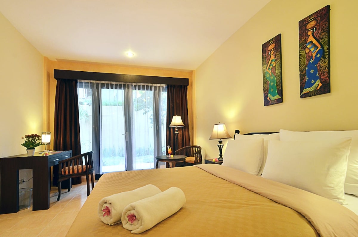 A spacious room features a comfortable bed with white linens, accompanied by two rolled towels placed at the foot. Elegant artwork adorns the walls, while large windows with sheer curtains overlook a private outdoor area. A desk with a lamp and plant adds functional decor.