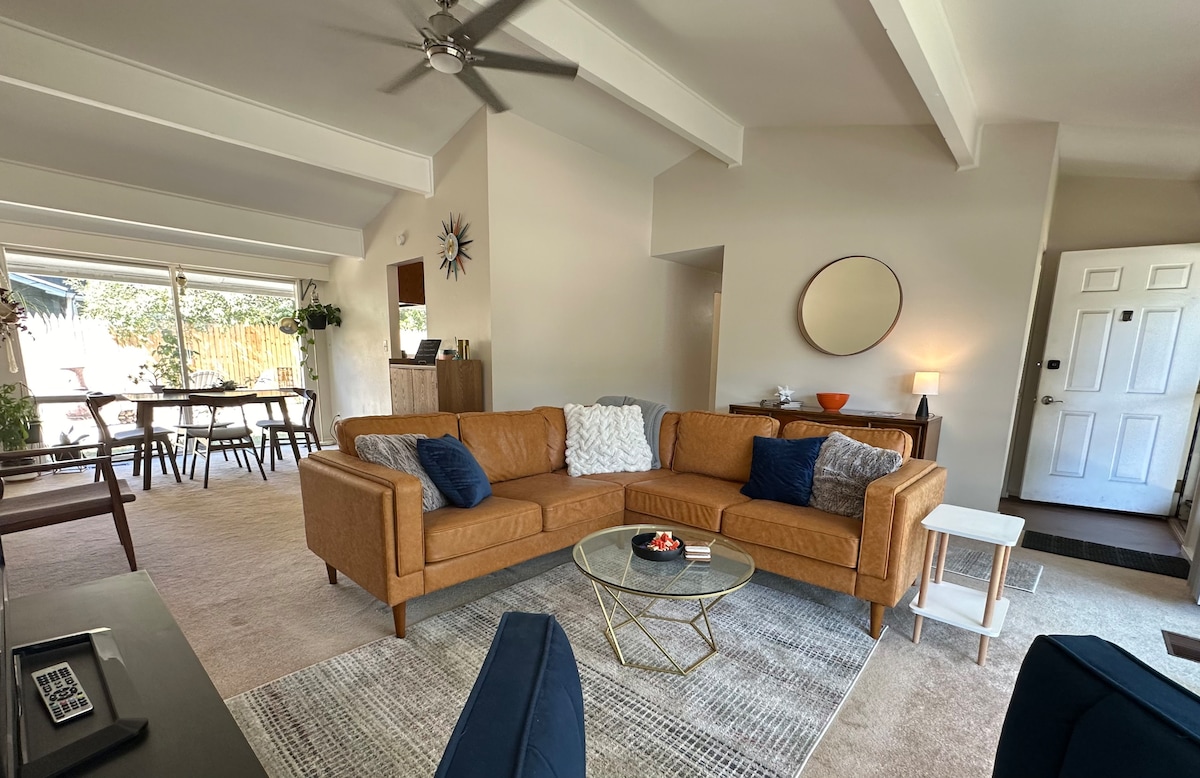A spacious living area features a mid-century modern sofa in warm tones, complemented by accent pillows. A glass coffee table sits at the center, with natural light streaming from large sliding doors that lead to the backyard. A decorative mirror and wall art enhance the welcoming atmosphere.