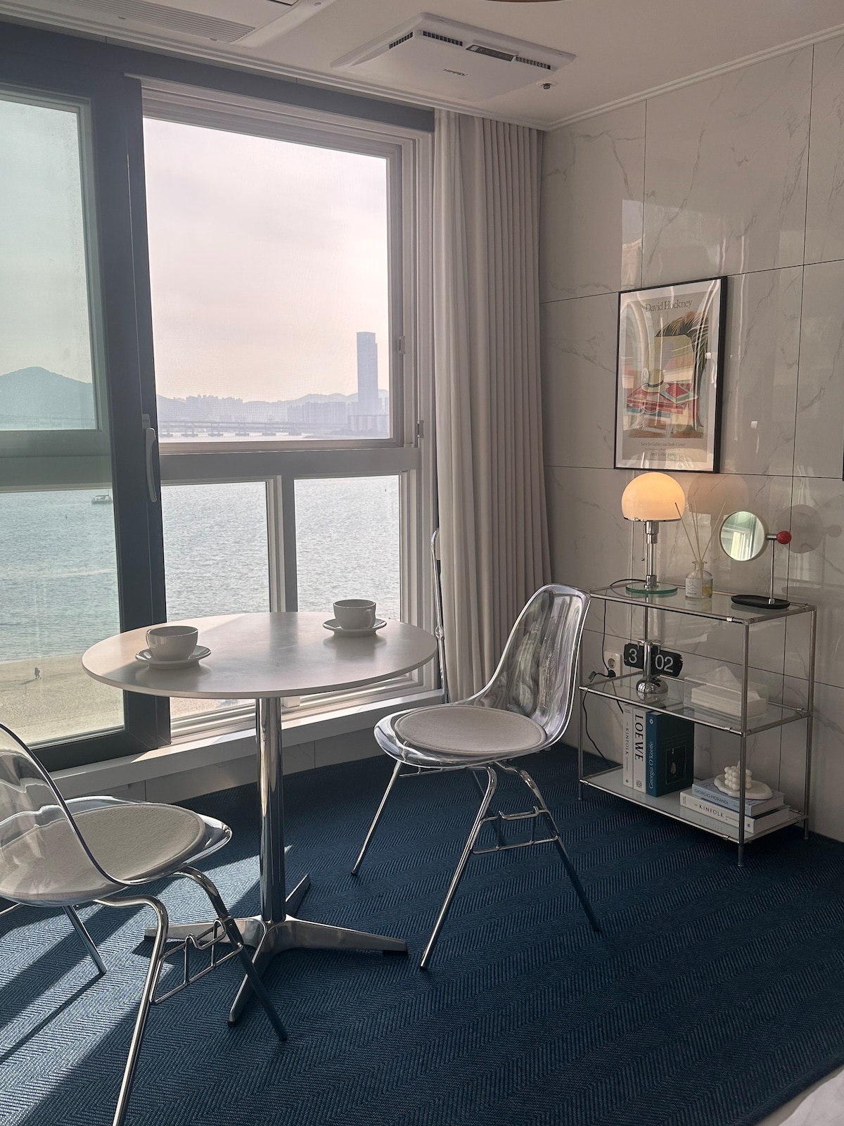 A small dining area features a round metallic table accompanied by two clear plastic chairs. Sunlight fills the space, reflecting off the large window with a view of the water and distant cityscape. A minimalist shelving unit with decor is positioned nearby.