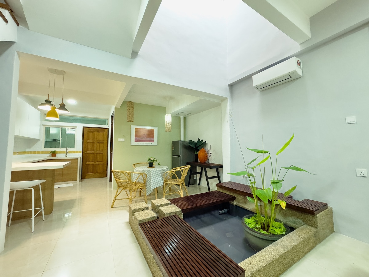An open living space is highlighted by a small indoor water feature surrounded by wooden decking. Lush greenery is present in a stone planter. A dining area with bamboo chairs and a light-colored table is visible, complemented by modern lighting fixtures above.
