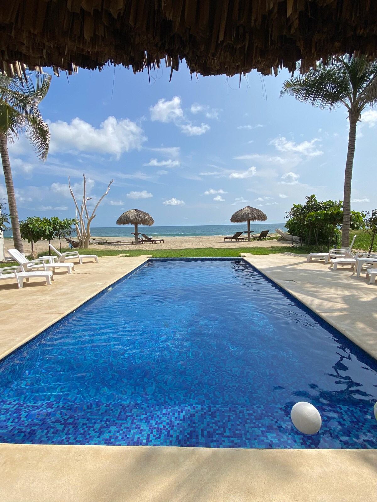 A refreshing swimming pool exhibits clear blue water, bordered by sun loungers. In the background, the sandy beach is framed by palm trees and thatched umbrellas, with the ocean visible under a bright sky adorned with scattered clouds.