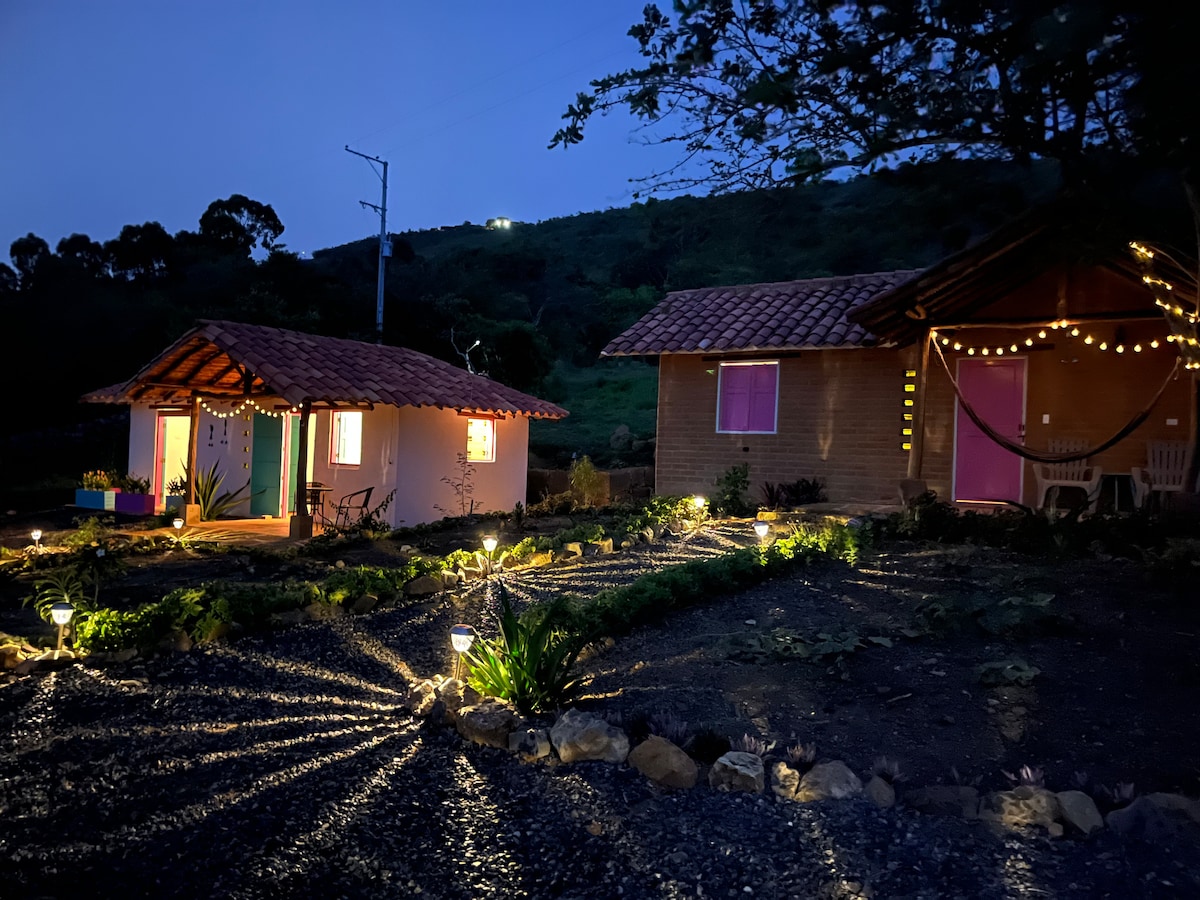 The exterior of the cabins is softly illuminated at night, showcasing their welcoming windows. Pathways are adorned with subtle lighting, leading to the inviting structures. A serene landscape with greenery surrounds the cabins, creating a peaceful ambiance under the evening sky.