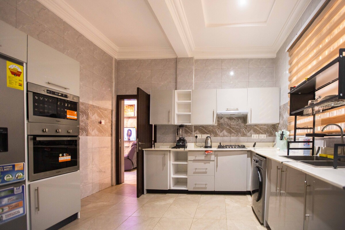 A modern kitchen features sleek grey cabinets and high-quality appliances, including an oven and a washing machine. A window with layered blinds allows natural light to fill the space, highlighting the tiled flooring and organized countertops.