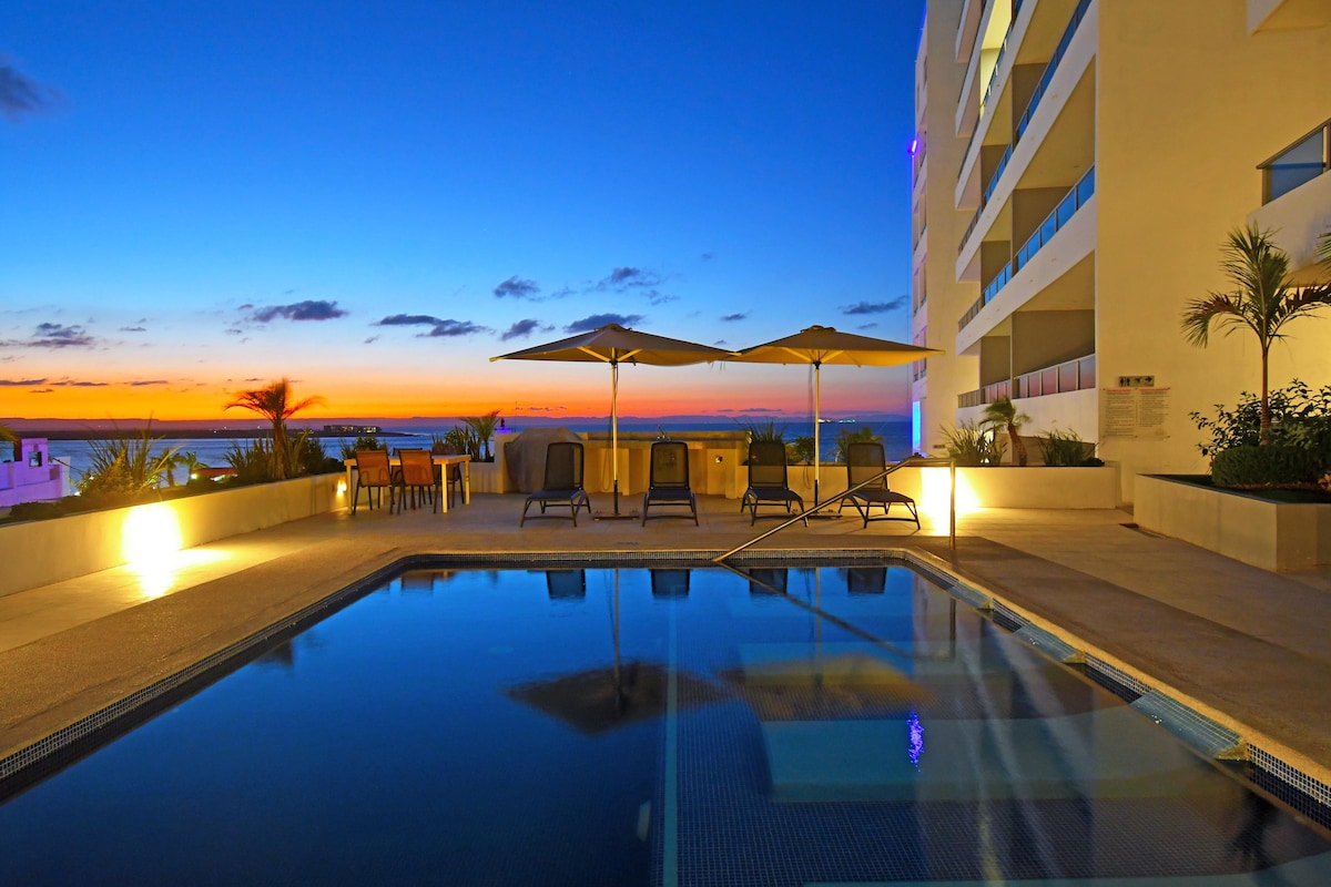 An inviting pool area is illuminated by soft lighting as the sun sets over the Sea of Cortez, creating a serene atmosphere. Lounge chairs and umbrellas provide comfortable seating options. The surrounding space is framed by tropical plants, enhancing the tranquil setting.