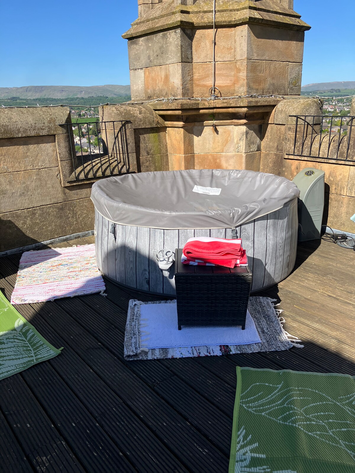 An inflatable hot tub is positioned on a wooden deck, surrounded by a scenic view of the landscape. Two textured mats lie nearby, with a small table displaying neatly arranged towels. The warm sunlight enhances the outdoor space, creating an inviting area for relaxation.