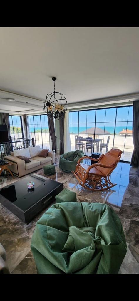 A bright living area features large glass windows offering views of the beach. Comfortable seating includes a couch and bean bags, complemented by a rocking chair. A dark coffee table sits atop polished flooring, with a dining table visible in the distance.
