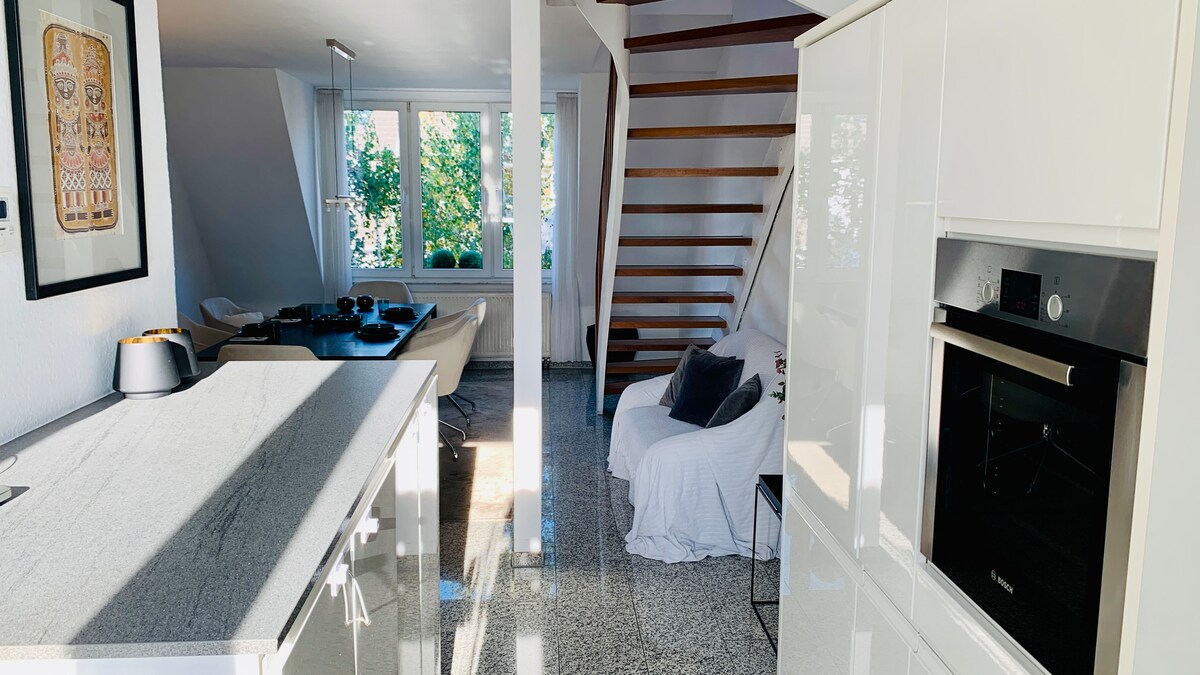 The open living space features a modern kitchen island with a sleek countertop, leading to a dining area with a light-colored table and chairs. Large windows allow natural light to illuminate the area, while a staircase with wooden steps enhances the overall design.